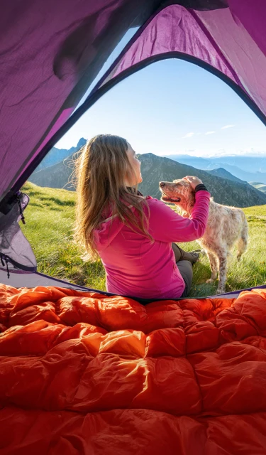 Femme assise dans une tente en montagne, caressant son chien tout en admirant le paysage